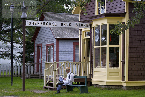 Ein kostümierten Mann sitzt außerhalb der Sherbrooke Drogerie entlang der Hauptstraße  Wharf Road  in Sherbrooke Dorfmuseum (eine restaurierte 1860 Stampfender Schiffbau Gemeinschaft und) in der Stadt von Sherbrooke entlang dem Marine-Antrieb  Highway 7  Nova Scotia  Kanada.