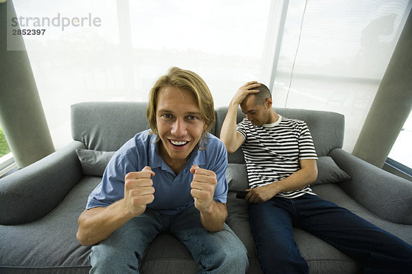 Zwei Männer  die auf dem Sofa sitzen und fernsehen  einer jubelt  der andere hält den Kopf.