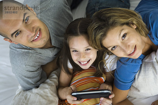 Mädchen liegt zwischen den Eltern  spielt Handheld-Videospiel  alle lächeln vor der Kamera