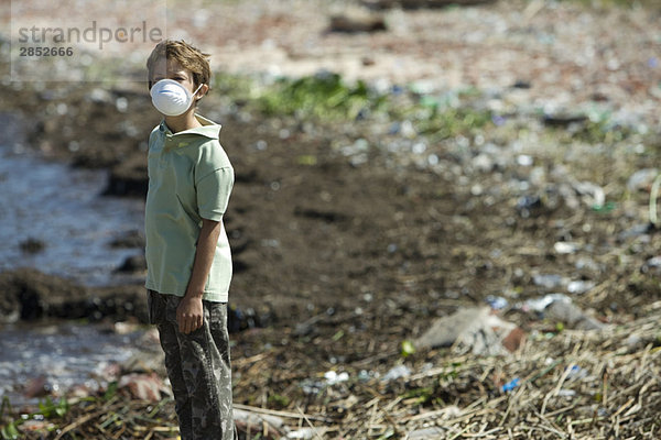 Junge steht am verschmutzten Ufer und trägt eine Verschmutzungsmaske.