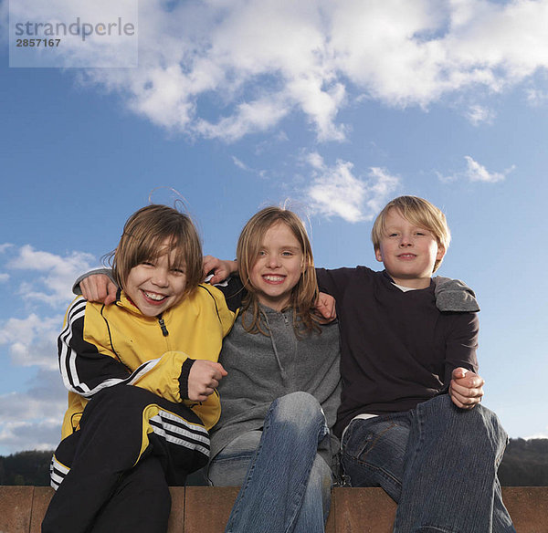 3 Kinder sitzen auf niedriger Wand  lächelnd