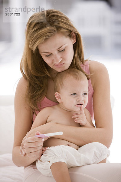 Frau betrachtet einen Schwangerschaftsteststab mit einer Tochter  die auf ihrem Schoß sitzt.