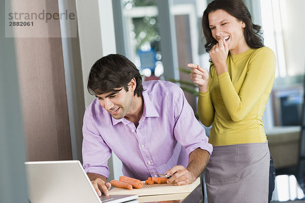 Mann kocht mit dem Rezept auf einem Laptop und eine Frau lacht ihn aus.