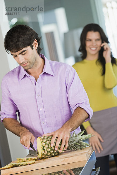 Ein Mann schneidet eine Ananas und eine Frau spricht auf einem Handy hinter ihm.