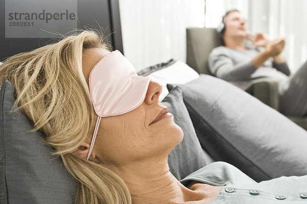 Frau mit Augenmaske und ihr Mann hört im Hintergrund Musik.