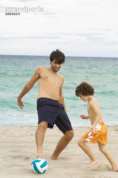 Junge spielt Fußball mit seinem Vater am Strand.