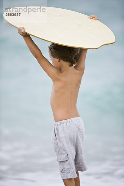 Rückansicht eines Jungen  der ein Bodyboard über den Kopf hält.