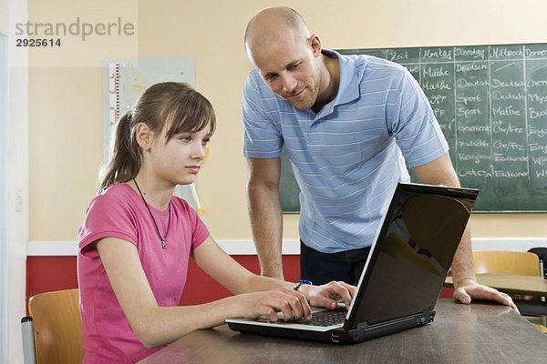 Ein Schulmädchen mit einem Laptop und einem Lehrer  der neben ihm steht.