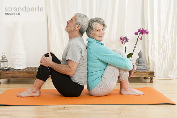 Ein älterer Mann und eine ältere Frau sitzen Rücken an Rücken auf einer Trainingsmatte.