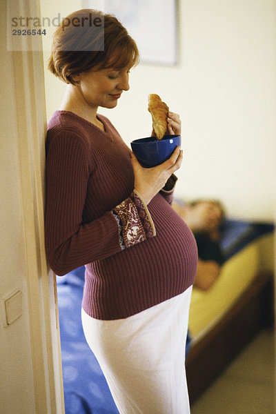Schwangere Frau  die sich in der Tür lehnt und Croissant isst.