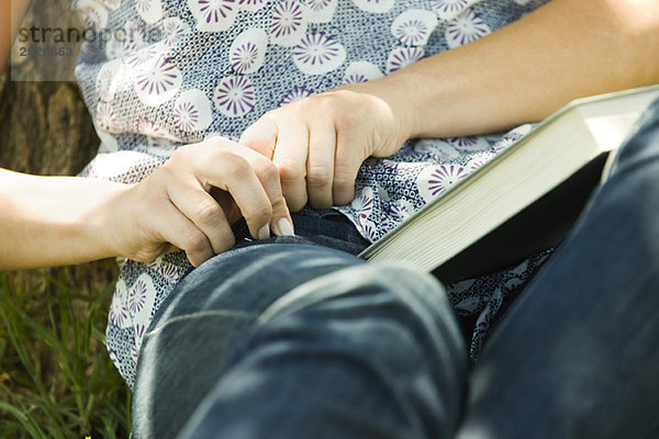 Junge Frau sitzt auf dem Boden mit Buch auf dem Schoß und greift nach etwas in der Tasche.