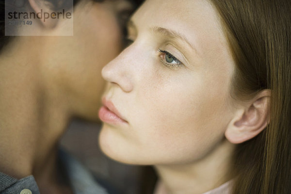 Junge Frau schaut weit weg  während der Mann ihre Wange küsst.