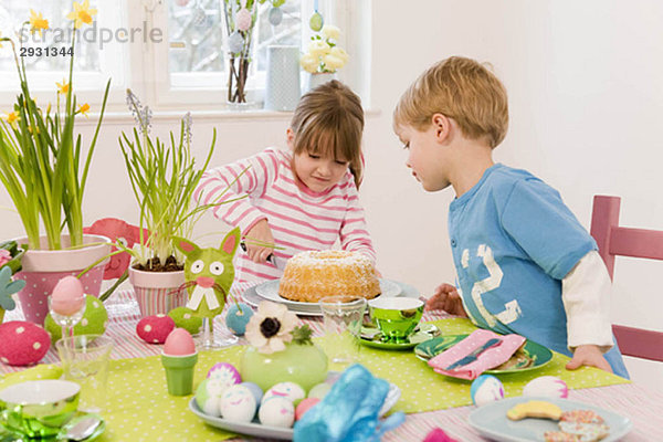 Mädchen und Junge schneiden Kuchen Ostertisch