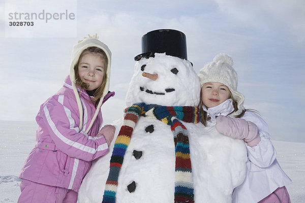 Zwei Mädchen (4-5) (8-9) neben Schneemann stehend  lächelnd  Portrait