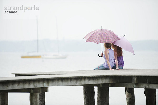 Zwei Frauen sitzen auf dem Steg  halten Regenschirm  Rückansicht