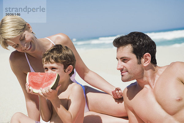 Junge isst eine Wassermelone mit seinen Eltern am Strand.