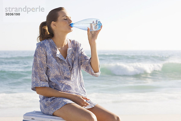 Frau sitzt auf einer Eisbox und trinkt Wasser am Strand.