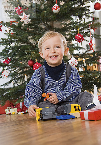 Kleiner Junge sitzt mit Spielzeug vor Weihnachtsbaum  lacht