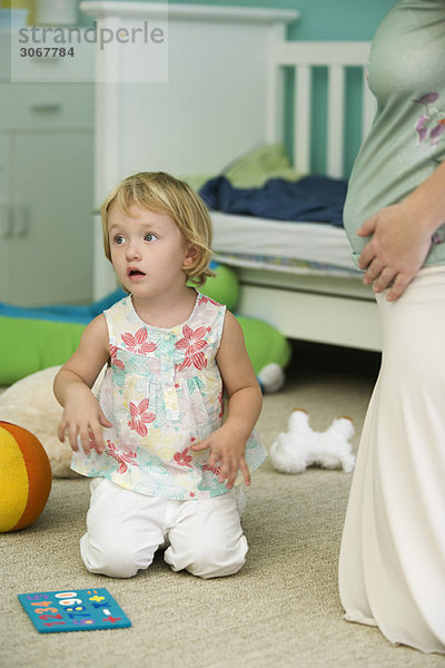 Kleines Mädchen kniend auf dem Boden im Kinderzimmer neben der schwangeren Mutter  abgeschnitten