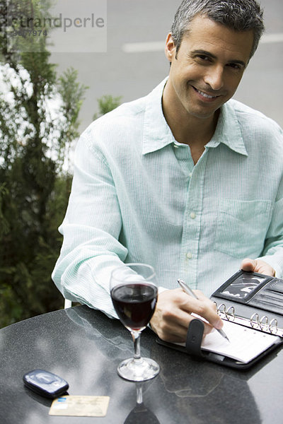 Mann sitzt am Kaffeetisch im Freien mit einem Glas Wein und schreibt in die Tagesordnung.