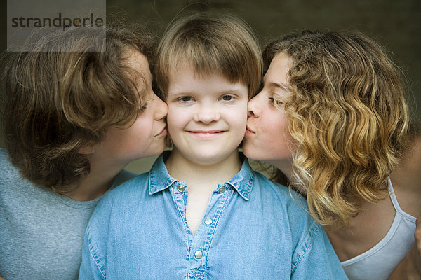 Mädchen mit Down-Syndrom lächelt vor der Kamera als zwei Schwestern ihre Wangen küssen  Portrait