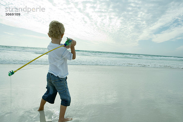 Junge hält Angelrute über Schulter am Strand  Rückansicht