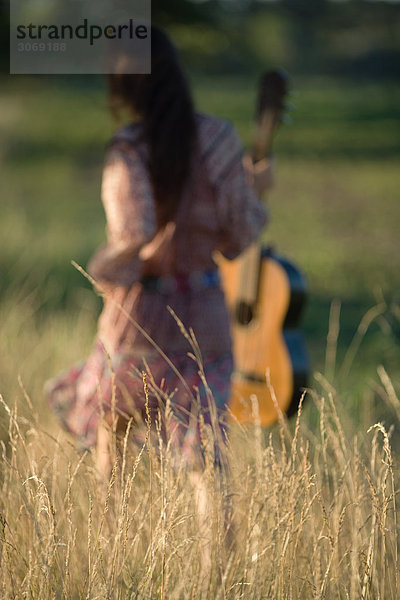 Rückansicht der Frau  die durchs Feld geht  Gitarre trägt  Fokus auf hohes Gras im Vordergrund
