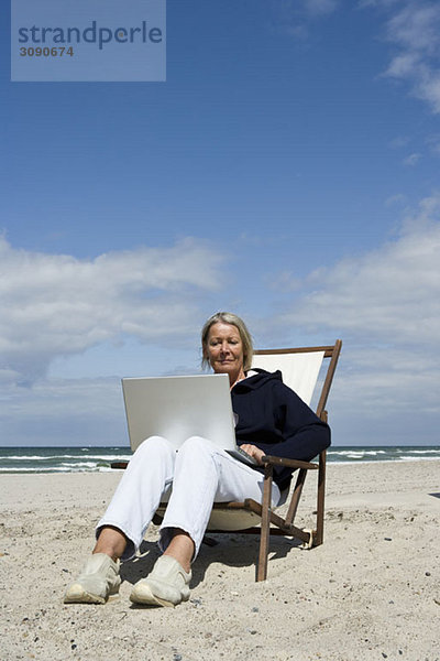 Eine ältere Frau sitzt in einem Sessel am Strand und benutzt einen Laptop.