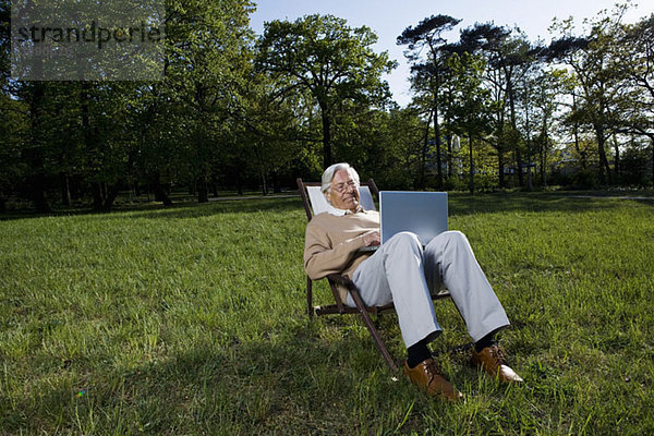 Ein älterer Mann  der in einem Sessel sitzt und einen Laptop benutzt  im Freien.