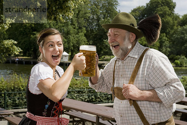 Ein traditionell gekleideter deutscher Mann und eine deutsche Frau in einem Biergarten mit Toastgläsern.