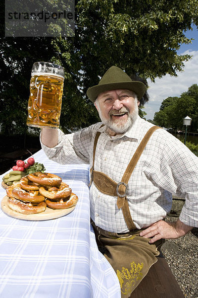 Ein traditionell gekleideter deutscher Mann in einem Biergarten  der sein Bierglas in Toast hebt.