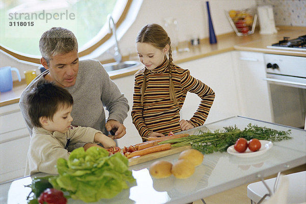 Vater mit Kindern in der Küche  hilft dem kleinen Sohn beim Gemüse schneiden