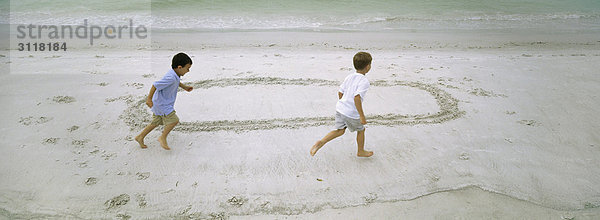 Jungen  die am Strand rennen und sich gegenseitig um den im Sand gezogenen Kreis jagen.