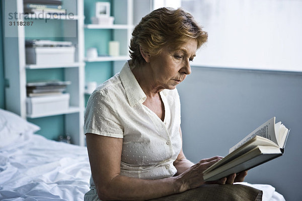 Seniorin auf dem Bett sitzend Lesebuch