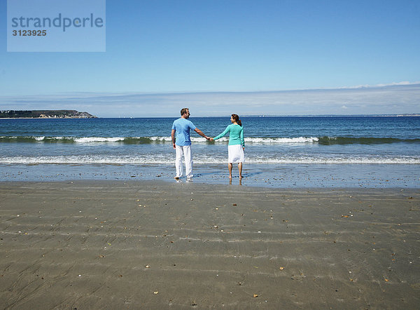 Paar am Strand stehend  Hände haltend  Rückansicht
