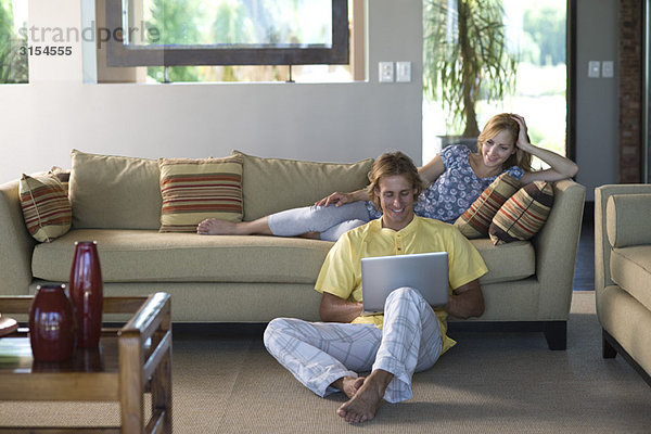 Paar im Wohnzimmer  Frau auf dem Sofa liegend  Mann auf dem Boden sitzend mit Notebook