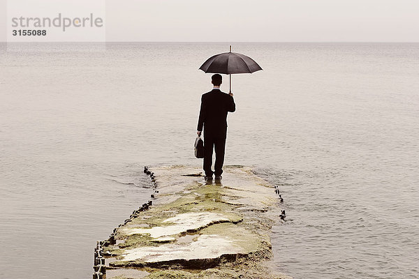 Man in suit holding umbrella and briefcase  standing at end of sinking pier