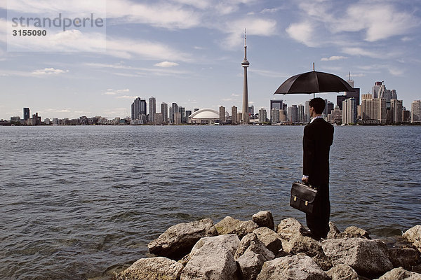Man in suit holding umbrella and briefcase  on breakwater with skyline in background  Toronto Islands  Ontario