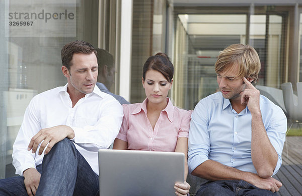 Deutschland  Köln  Drei Geschäftsleute sitzen Seite an Seite  mit Laptop