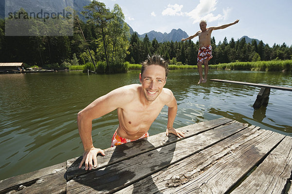 Italien  Südtirol  Mann im Vordergrund am Steg  Senior im Hintergrund springt in den See  Portrait