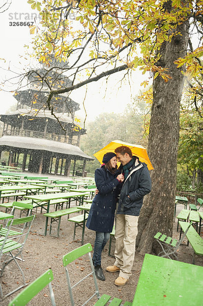 Deutschland  Bayern  München  Englischer Garten  Paar im verregneten Biergarten mit Regenschirm  Chinesischer Turm im Hintergrund  Portrait