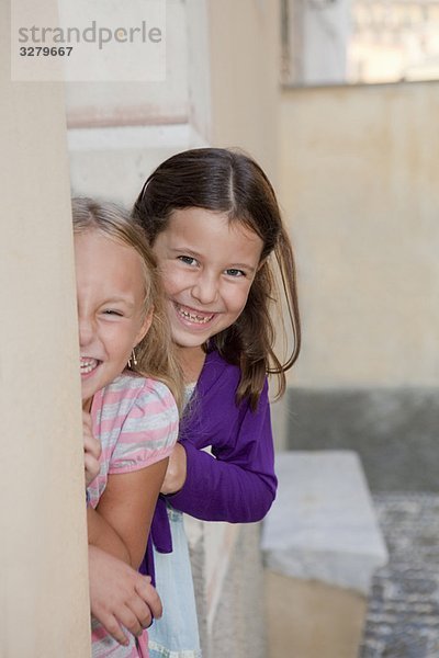 zwei Mädchen  die sich hinter der Wand verstecken und lachen.