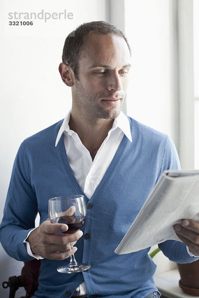 Ein Mann  der ein Glas Wein trinkt und eine Zeitschrift liest.