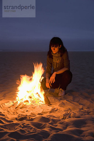 Eine Frau  die in ein Feuer starrt  das am Strand brennt.