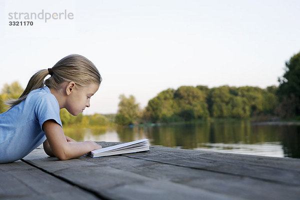 Ein junges Mädchen liest ein Buch auf einem Holzsteg.