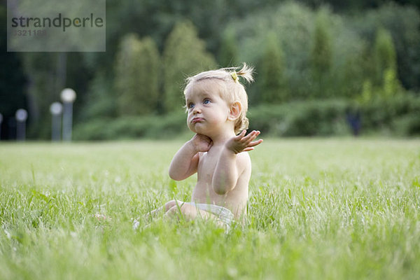 Ein kleines Mädchen  das auf Gras in einer Windel sitzt und verwirrt aussieht.