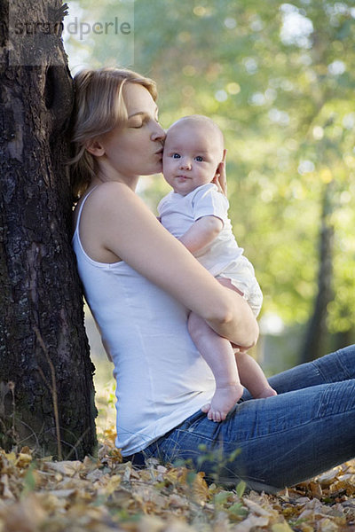 Eine Mutter sitzt an einem Baum und küsst ihr Baby.