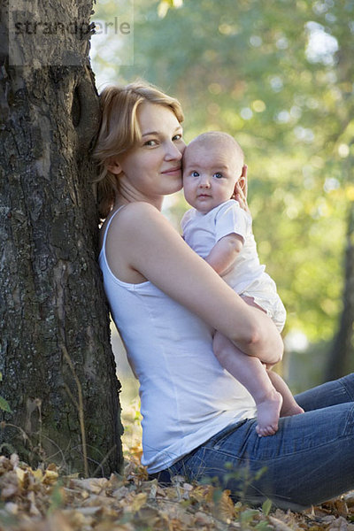 Eine Mutter sitzt an einem Baum  küsst ihr Baby und schaut in die Kamera.