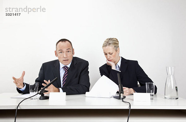 Ein Mann und eine Frau sitzen an einem Schreibtisch mit Mikrofonen und Dokumenten.