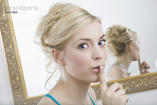 Eine Frau mit dem Finger auf den Lippen vor dem Spiegel  mit Blick auf die Kamera.